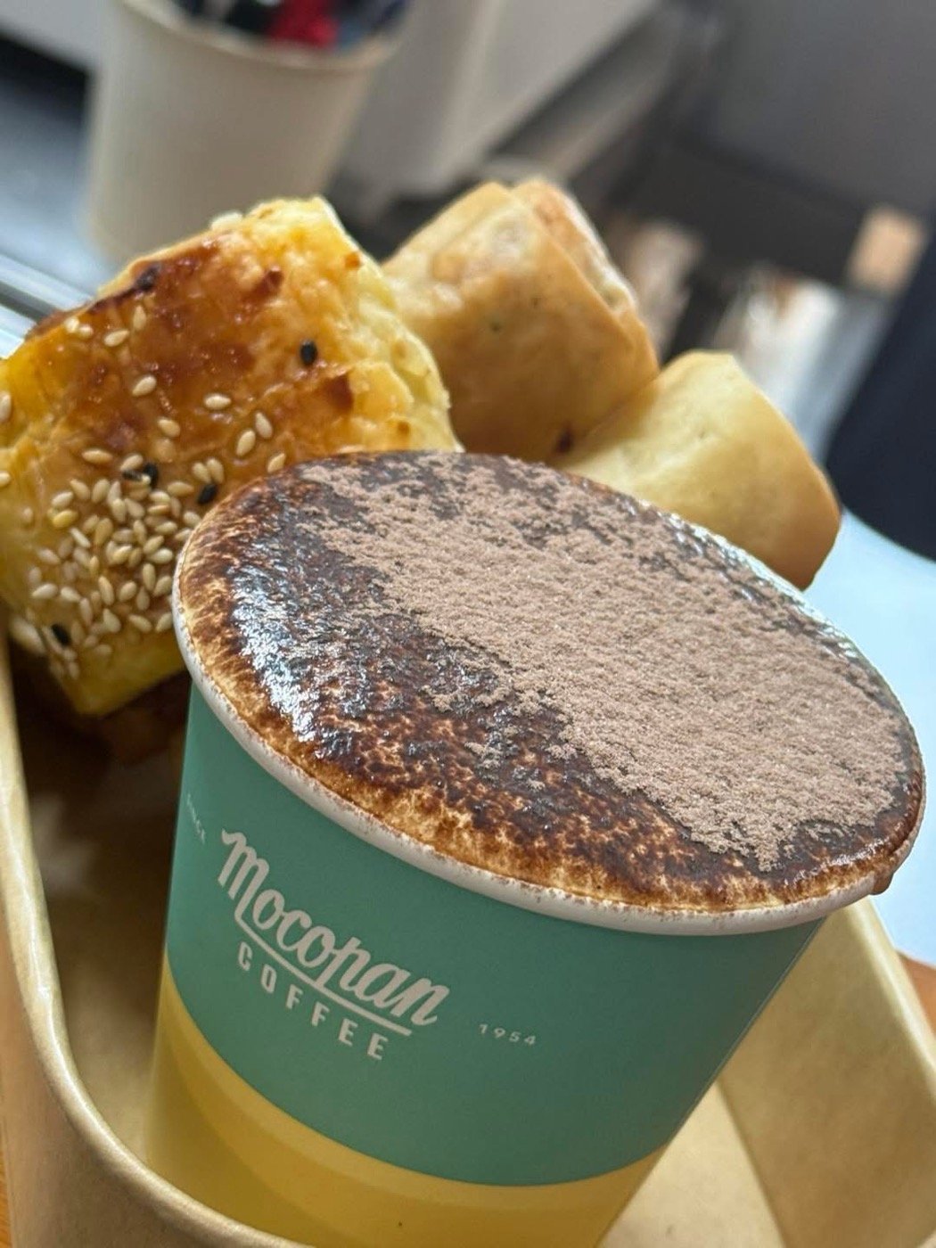 A takeaway cappuccino dusted with chocolate beside golden sesame-topped pastries on a tray