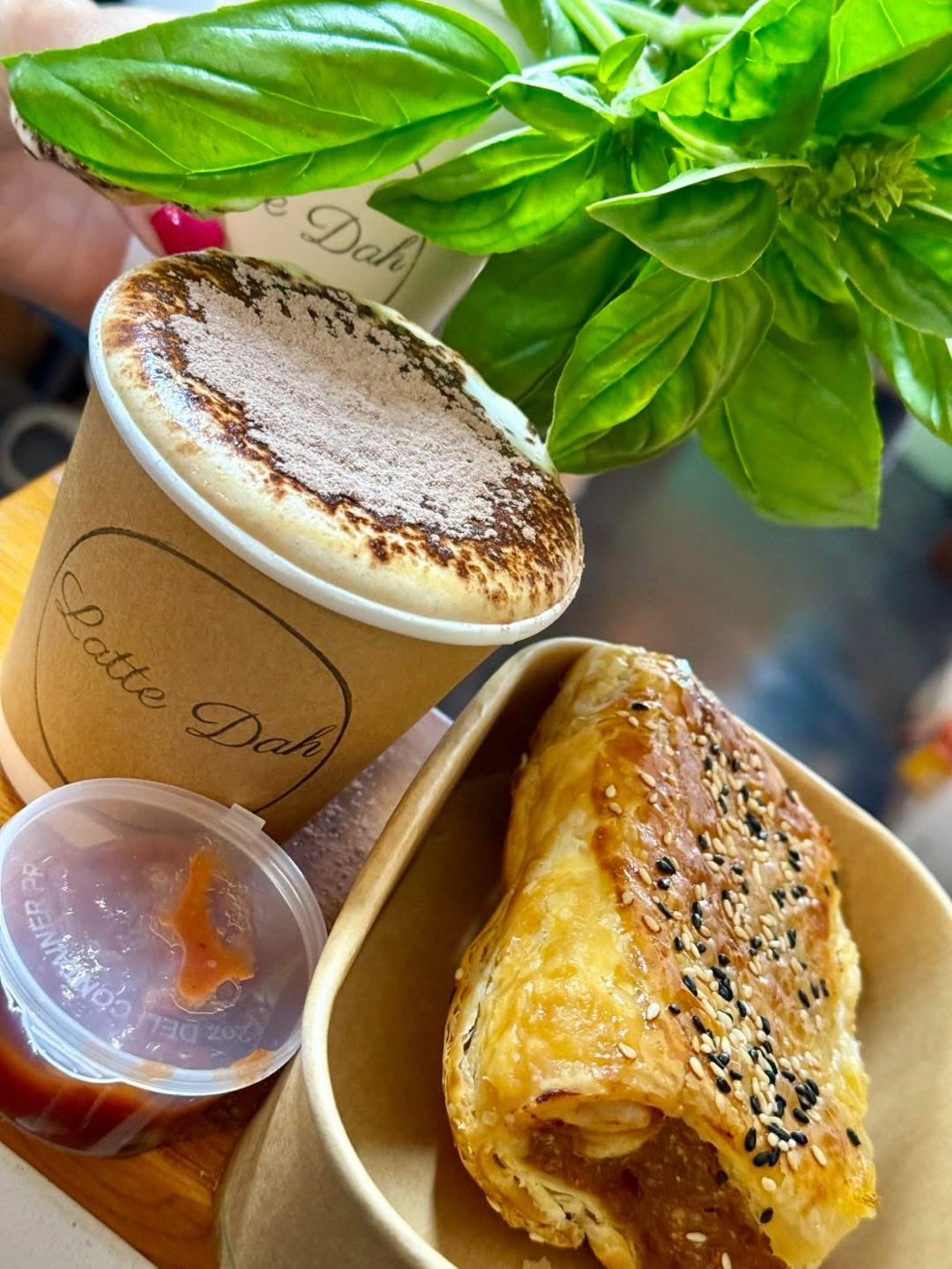 A Latte Dah takeaway cup beside a sesame-topped sausage roll in a paper tray, with a pot of relish and fresh basil leaves