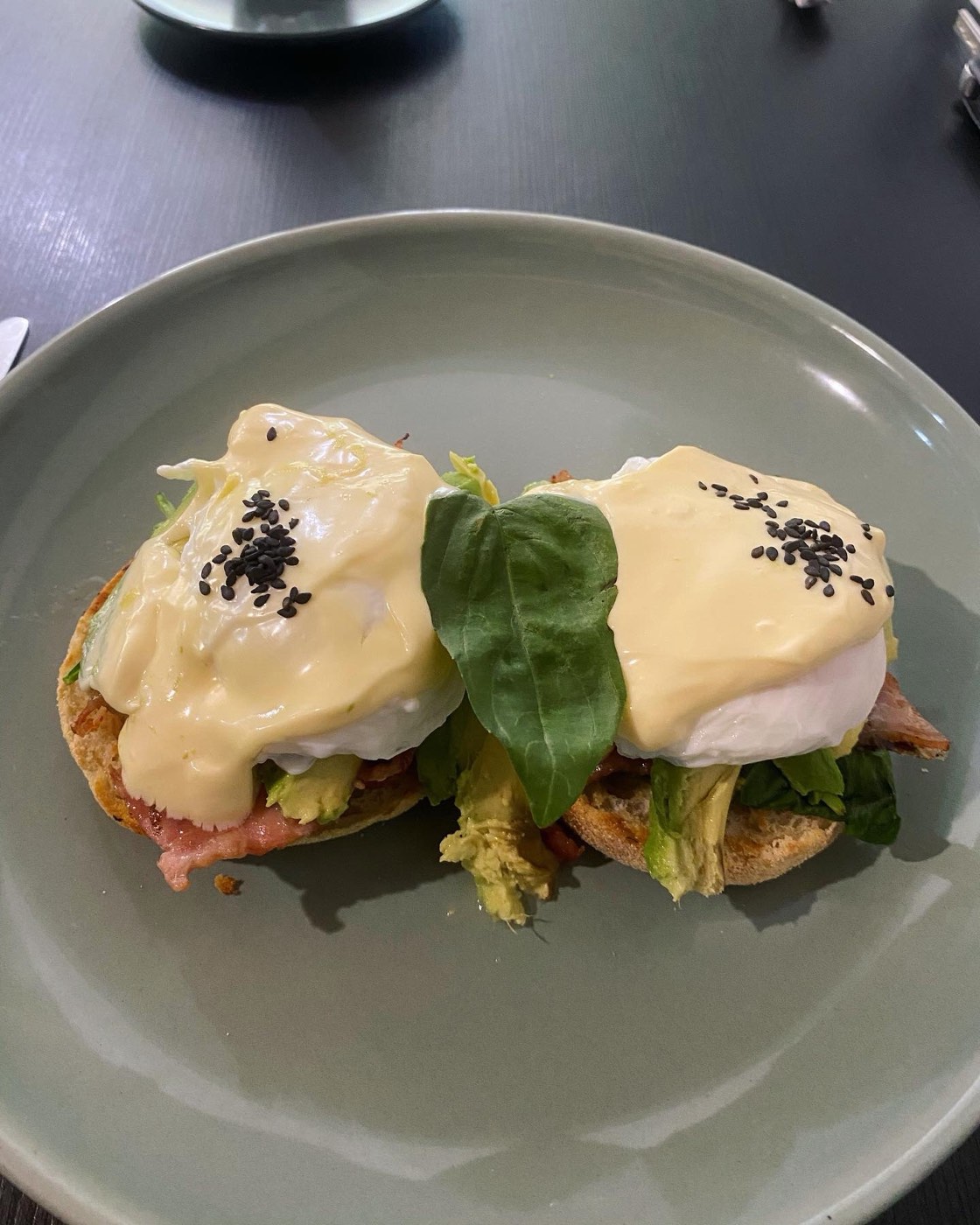 Two eggs benedict with hollandaise on English muffins with avocado, bacon and a basil leaf, on a grey plate