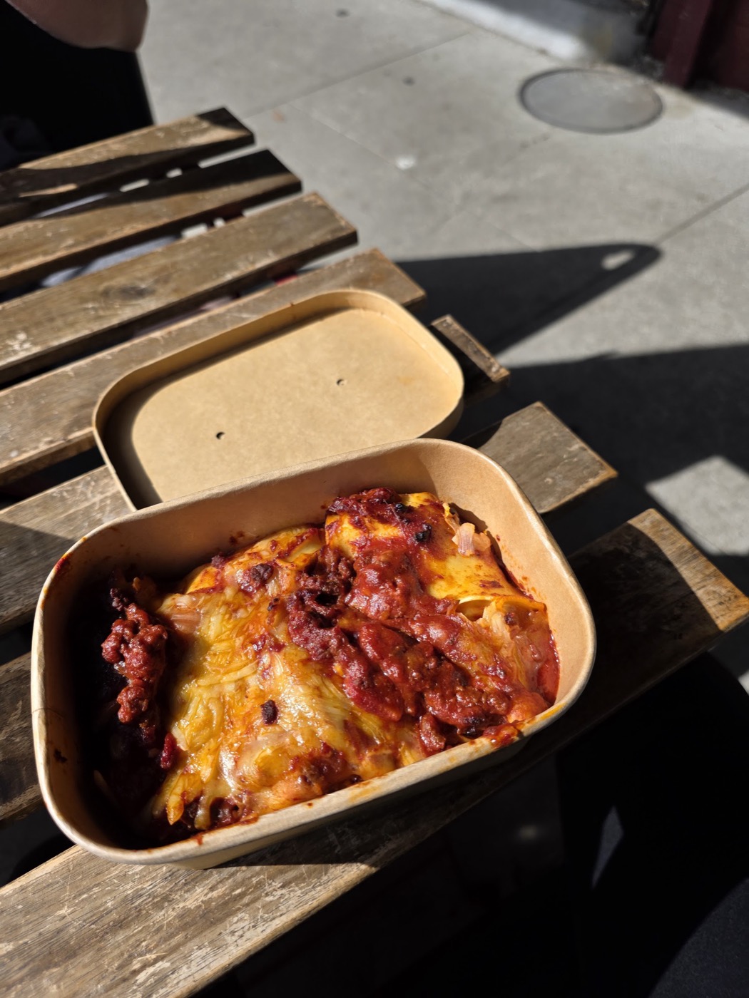 A takeaway container of baked lasagne topped with melted cheese, on a wooden bench