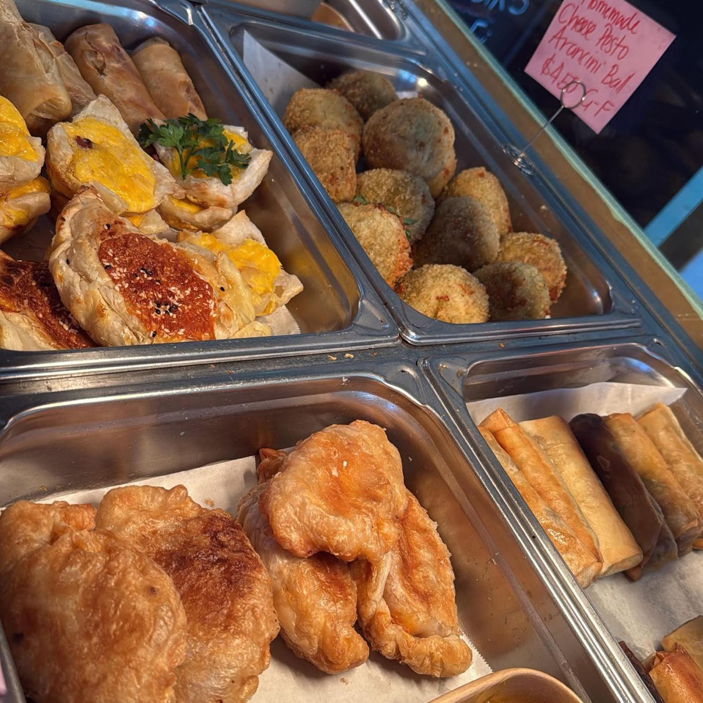 Steel trays in a hot display holding spring rolls, arancini balls, pasties and golden fried pastries